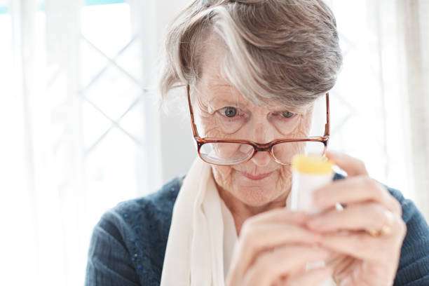 An aged woman peers over her spectacles trying to read the instructions for the medication she holds.